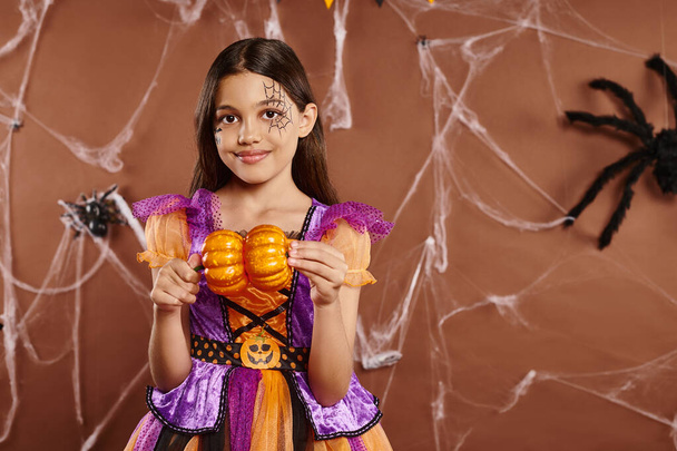 wesoła dziewczyna z makijażem pajęczyny w kostiumie Halloween trzymając dynie na brązowym tle - Zdjęcie, obraz