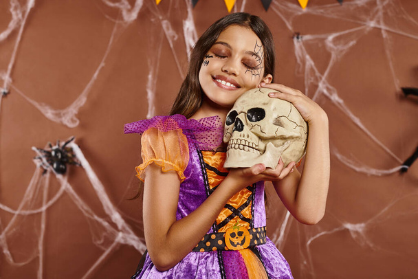 encantadora chica en vestido sosteniendo cráneo y sonriendo sobre fondo marrón, Halloween temporada espeluznante - Foto, Imagen