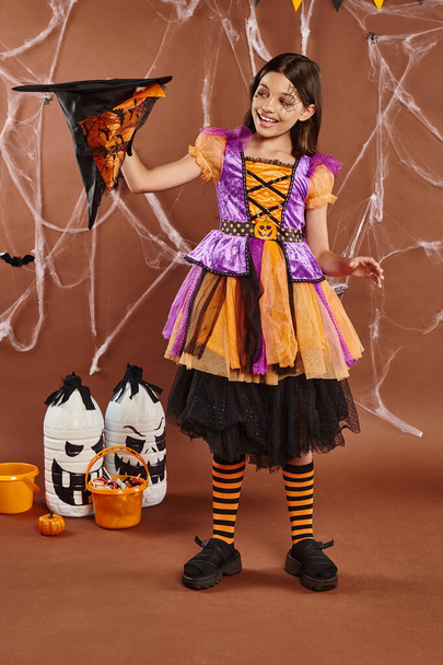 niño alegre en disfraz de bruja de Halloween sosteniendo sombrero apuntado al revés sobre fondo marrón - Foto, Imagen