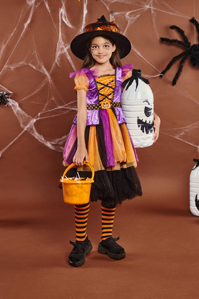 chica feliz en sombrero de bruja y Halloween traje celebración cubo con dulces y decoración espeluznante bricolaje - Foto, Imagen