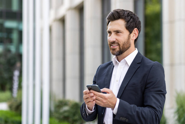 Close-up photo of a young businessman standing in a business suit and using a mobile phone, smiling and looking to the side. - Foto, Bild