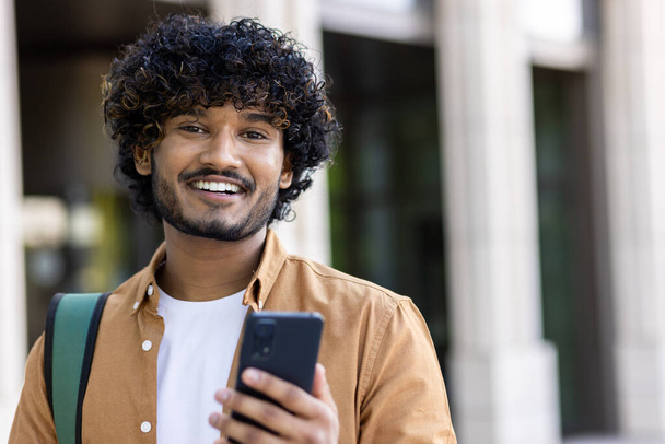 Close-up photo of a young Indian man smiling at the camera, holding a phone and a backpack. - Photo, image