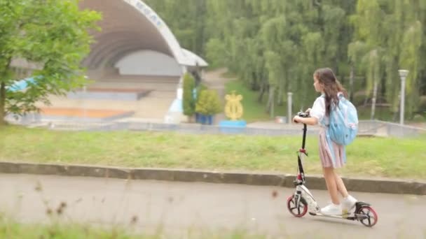 Une petite fille avec un sac à dos va à l'école en scooter. Vue arrière. Mode de vie d'enfance heureux. - Séquence, vidéo