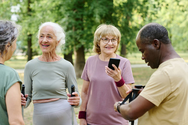 Группа пожилых людей собирается на улице, чтобы тренироваться, они разговаривают друг с другом и используют смартфон - Фото, изображение