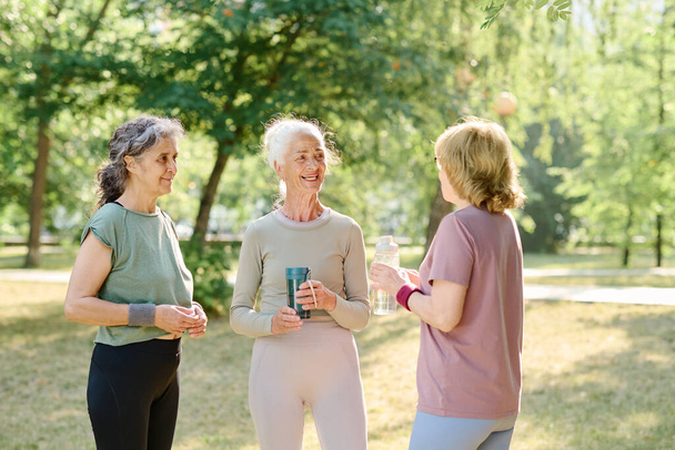 Seniorinnen treffen sich im Park zum Sporttraining, reden miteinander und trinken Wasser aus Flaschen - Foto, Bild