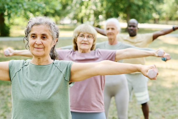 Grupo de personas mayores de pie en fila y haciendo ejercicio con pesas al aire libre - Foto, imagen