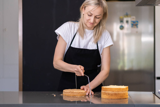Focused blonde housewife showing tutorial of cutting baked cake with string knife standing at big table food blogger in black apron doing confectionery training at home - Photo, Image