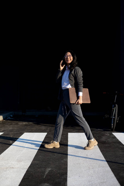 молодая азиатская деловая женщина идет и использует мобильный телефон в своих руках городской фон современного офисного здания. Привлекательная деловая женщина гуляет по улицам города с помощью смартфона. - Фото, изображение