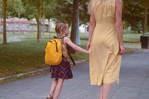 Een klein meisje, vrolijk hoppend, begeleidt haar moeder naar school door het park met rugzakken..  - Foto, afbeelding