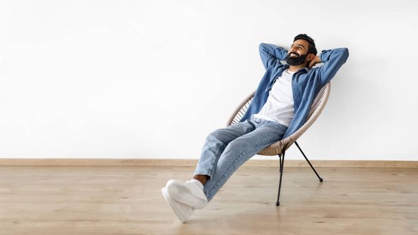 Hombre indio joven relajado relajándose con las manos detrás de la cabeza en el sillón interior, disfrutando de la comodidad y el descanso en la silla cerca de la pared del estudio blanco. Panorama con espacio vacío para publicidad de texto - Foto, Imagen