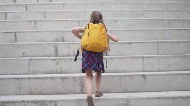 Malá holka se žlutou školní brašnou jde nahoru kamenné kroky v moderním městě školačka s batohem na vysokých schodech backside pohled dítě student jde do školy - Záběry, video