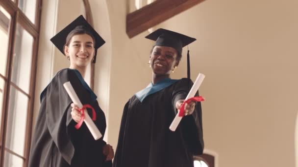 Low-angle-Slowmo-Porträt von zwei fröhlichen Studentinnen in Abschlusskleidern, die ihre Diplome mit roten Bändern in die Kamera halten und lächeln - Filmmaterial, Video