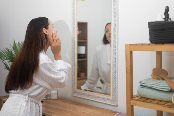 Νεαρή γυναίκα με ρόμπα κοιτάζοντας στον καθρέφτη στο μπάνιο όμορφη κυρία αγγίζοντας το δέρμα του προσώπου στέκεται δίπλα τουαλέτα στο σπίτι γυναίκα απολαμβάνοντας καθαρό δέρμα σε αντανάκλαση - Φωτογραφία, εικόνα