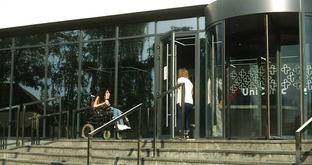 Frau mit körperlicher Behinderung fährt mit motorisiertem Rollstuhl in Klinik-Eingang. Medizinisches Personal, männliche Krankenschwester lernt die Patientin kennen und hilft ihr, hereinzukommen. Krankenhaus oder moderne medizinische Einrichtung im Freien. - Foto, Bild