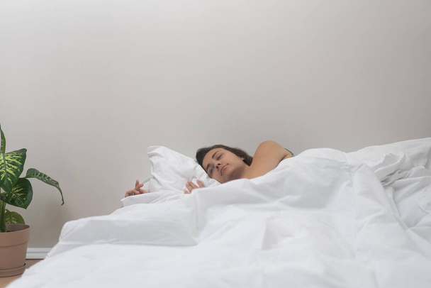 Uvolněná žena spí na posteli s lehkým lůžkoviny v jasném prostoru v blízkosti dekorativní květiny v květináči žena dostane energii ze spánku relaxační v moderním bytě - Fotografie, Obrázek