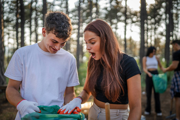 couple or teenage friends gen Z male and female caucasian man woman picking up waste garbage plastic bottles and paper from forest cleaning up nature in sunny day environmental care ecology concept - Photo, Image