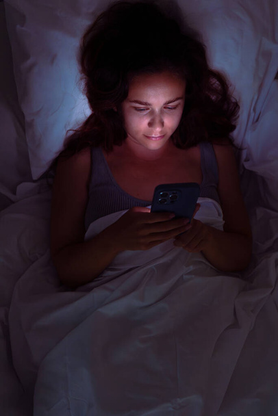 暗闇に包まれた女性は,ベッドの中で彼女の携帯電話を見つめ,不眠症とソーシャルメディアの執着の領域を垣間見る.  - 写真・画像