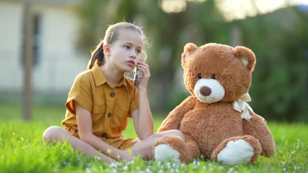 Ongelukkig kind meisje hebben een discussie tijdens een gesprek aan de telefoon met haar vriend terwijl zitten met een teddybeer buiten op groen gras gazon. Begrip vriendschapsproblemen. - Video