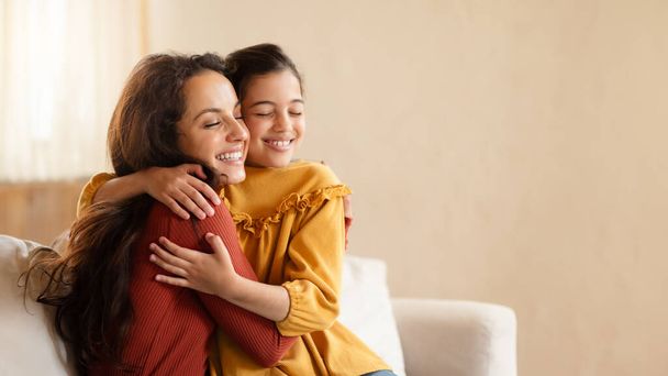 Cheerful cute hispanic preteen daughter girl hugging millennial mom, expressing their love and enjoying spare time in modern living room interior. Panorama, free space. Mothers support and care - 写真・画像