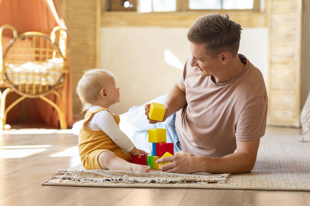 Glücklicher junger Vater, der mit seinem kleinen Baby-Sohn oder seiner Tochter zu Hause spielt, lächelnder Vater und süßes Kleinkind, das Bausteine stapelt, während es sich im Wohnzimmer auf dem Boden entspannt, Kopierraum - Foto, Bild