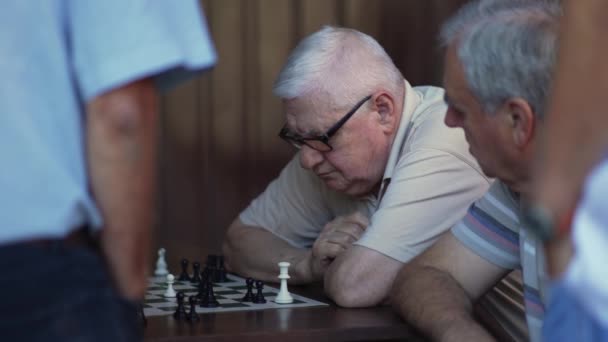 Valkoihoinen vanha harmaatukkainen mies istuu pöydässä shakkilaudan edessä ulkona.. - Materiaali, video