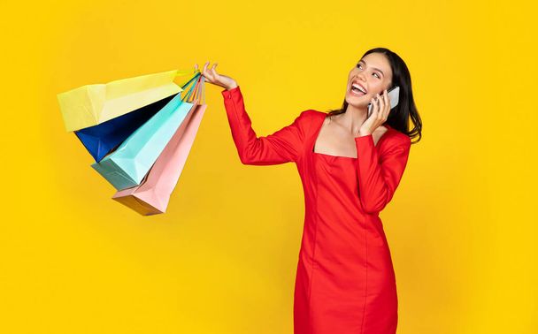 Portret van een lachende jonge vrouw die praat op een mobiele telefoon en boodschappentassen vasthoudt, vrolijke winkelverslaafde dame die haar vriendin belt, verkoopnieuws deelt, op een gele studio achtergrond staat, kopieerruimte - Foto, afbeelding
