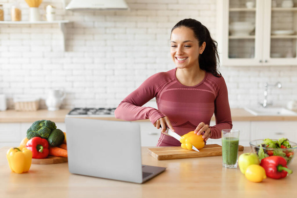 Cheerful fitness lady looking at laptop cooking vegetable salad, cutting bell pepper, preparing healthy meal for dinner using website, standing at kitchen table indoor. Nutrition And slimming - Фото, изображение