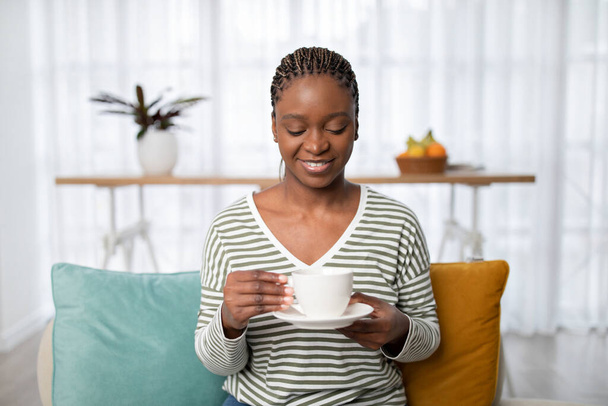 Портрет веселой расслабленной красивой молодой черной женщины в удобной домашней одежде, сидящей на диване, держа чашку белого фарфора и блюдце, наслаждаясь свежим ароматным кофе дома, копировать пространство - Фото, изображение