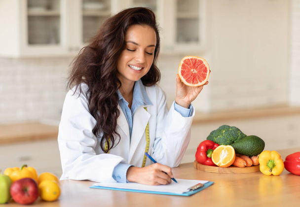 Happy caucasian woman doctor nutritionist making notes of diet plan at table with fruits, holding grapefruit, free space. Work, professional weight loss and health care - Photo, Image