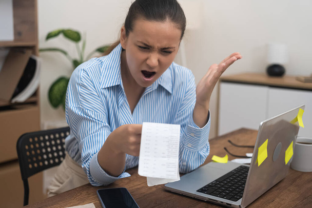 Stres finansowy: zestresowana kobieta, siedząca przy biurku w swojej siedzibie, gapiąca się na papierowe rachunki, oszołomiona wysokimi stawkami płatności.  - Zdjęcie, obraz