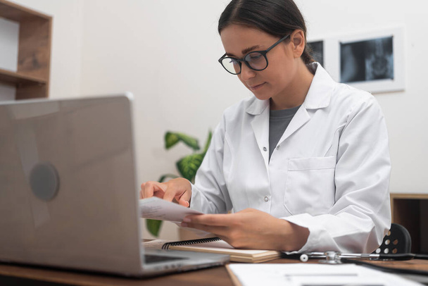 Lady doctor in glasses reads results of patient tests working at desk with laptop and documents pleasant young woman makes diagnosis working in modern medical clinic - Photo, Image