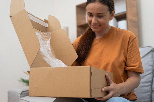 Чудова доставка: Зміст жінки на дивані відкриває пакет, демонструючи своє щастя після онлайн-шопінгу з експрес-доставкою.  - Фото, зображення