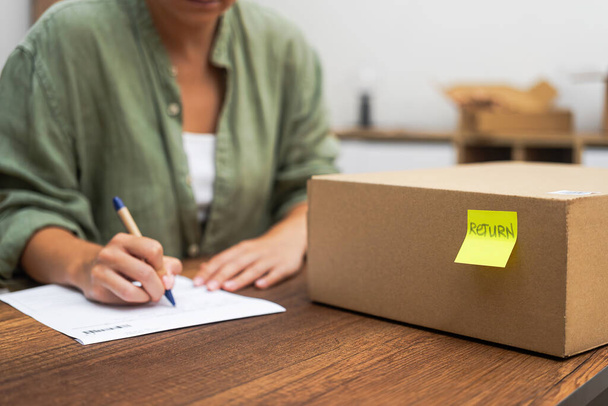 Female writes complaint about delivery near parcel with yellow sticker with word Return buyer dissatisfied with received box of goods with desire to send back - Photo, Image