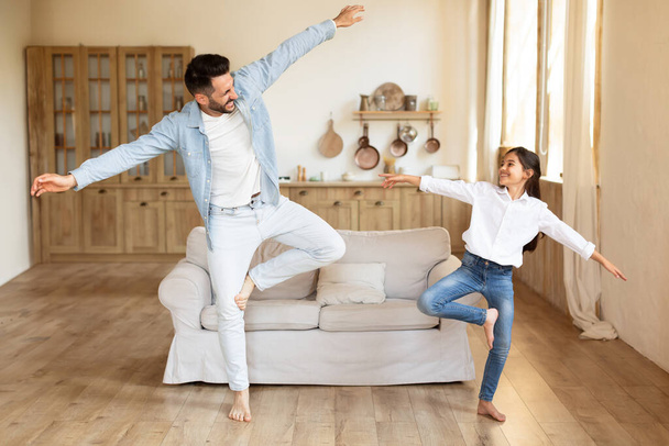 Joyful daddy and his daughter girl dancing to music and doing funny moves together while fooling at home. Father enjoying time with kid, having fun on weekend in living room indoors, full length - Photo, Image