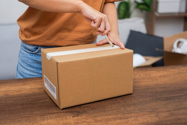 Housewife opens cardboard parcel with ordered goods with stationery knife on wooden table unpacking box on blurred background of open box and plant in pot in premise - Photo, Image