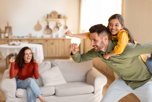 Family Fun And Playtime. Funny Dad Piggybacking And Riding His Joyful Daughter While Mom Looking At Them, Laughing And Playing Together At Home In Living Room Interior. Selective Focus - Photo, Image