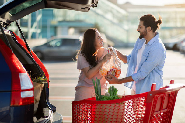 Щаслива латинська пара стоїть на парковці і завантажує паперові пакети після покупки у відкритий багажник. Люблячий чоловік і жінка роблять продукти разом, кладуть речі в авто - Фото, зображення