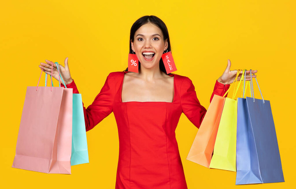 Портрет счастливой молодой женщины, держащей сумки для покупок и носящей ярлыки в качестве серьг, веселой красивой продавщицы, наслаждающейся сезонными продажами и скидками, позирующей изолированной на жёлтом фоне - Фото, изображение