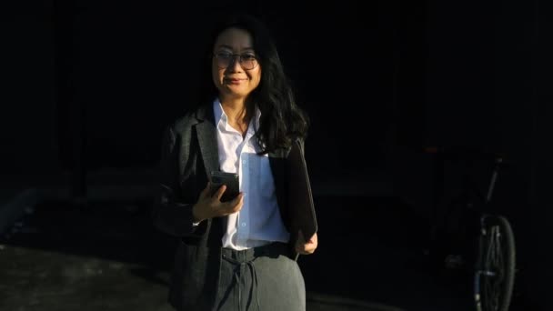 Mujer asiática sonriente usando traje formal usando el teléfono móvil escribiendo mensajes de texto caminando sosteniendo portátil fuera de la ciudad de teléfonos celulares de negocios smartphone cámara lenta - Metraje, vídeo
