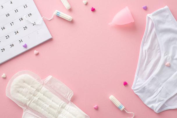 Horní pohled na předměty menstruační péče, jako je podložka, tampóny, menstruační pohár, kalhotky, kalendář, srdce, uspořádány na pastelově růžovém povrchu s rámečkem pro text nebo reklamu - Fotografie, Obrázek