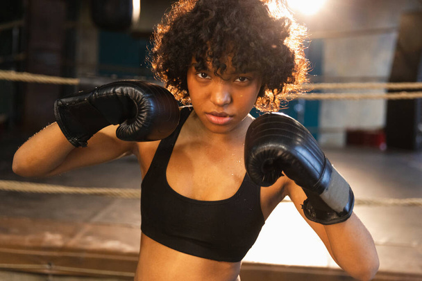 Bağımsız kız gücü diye bağır. Boks eldivenli kızgın Afrikalı Amerikalı kadın dövüşçü boks ringinde oldukça agresif görünüyor. Güçlü savaşçı kız yumrukları eğitiyor. - Fotoğraf, Görsel