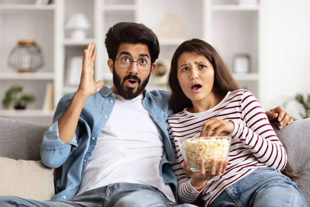 Schockiertes hinduistisches Millennial-Paar, das zu Hause auf der Couch sitzt, fernsieht, triller. Liebender junger Mann und Frau mit schockiertem Gesichtsausdruck, der auf den Bildschirm schaut und Popcorn isst - Foto, Bild