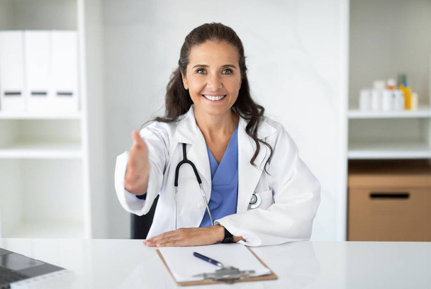 Ευτυχισμένο όμορφο φιλικό ώριμη γυναίκα γιατρός που έχει τη δική της πρακτική. Χαμογελώντας Ευρωπαίος ιατρός ή καρδιολόγος με στολή λευκού εργαστηρίου κάθεται στο γραφείο στο σύγχρονο γραφείο της, απλώνοντας το χέρι - Φωτογραφία, εικόνα