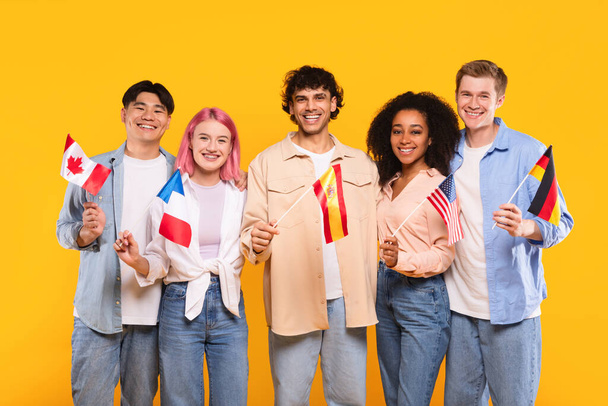 Internationale Freundschaft. Glückliche multiethnische Menschen, die verschiedene Länderfahnen in der Hand halten und in die Kamera lächeln, stehen auf gelbem Studiohintergrund. Moderne Ausbildung von Studenten oder Einwanderern - Foto, Bild