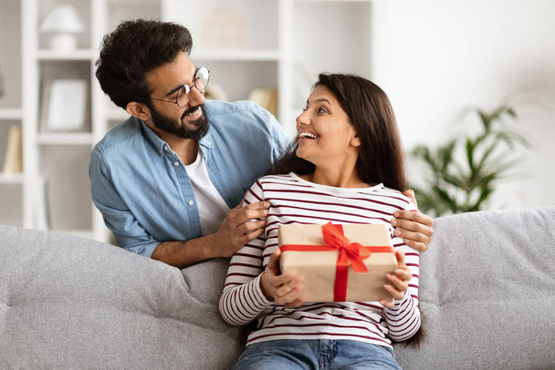 Kochający wschodni mężczyzna witający swoją podekscytowaną żonę z rocznicą ślubu, dający pudełko prezentów. Wesoły piękny młody Indianin para świętuje urodziny razem w domu - Zdjęcie, obraz
