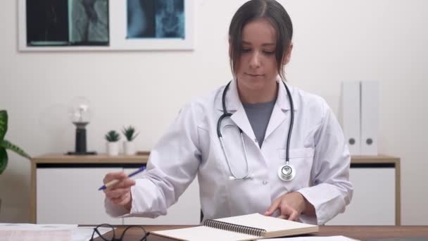 Thérapeute examine les résultats des tests du patient assis au bureau femme médecin avec stéthoscope tient presse-papiers avec des documents assis dans le bureau avec des images de rayons X sur le mur - Séquence, vidéo