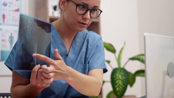 γιατρός με μπλε στολή που δείχνει με το δάχτυλο σε ακτινογραφία φιλμ που δείχνει στον ασθενή σε φορητό υπολογιστή κατά τη διάρκεια της online τηλεϊατρικής διαβούλευσης - Πλάνα, βίντεο