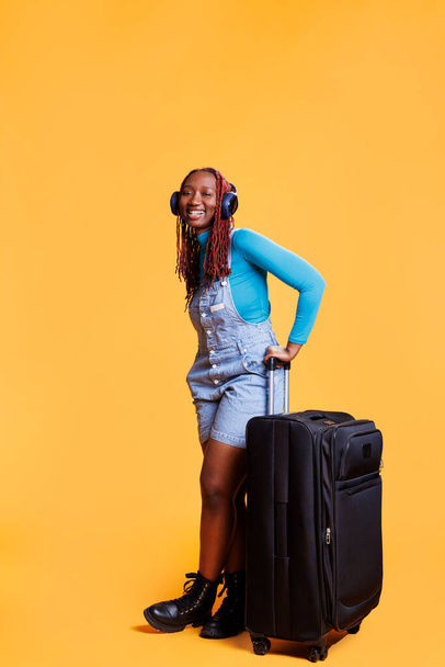 Young person travelling on vacation, carrying trolley bag luggage in studio and listening to music. Happy traveller feeling excited about international voyage destination, wearing headset. - Photo, Image