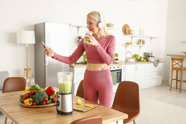 Donna matura sportiva con cellulare e bicchiere di frullato sano in cucina - Foto, immagini