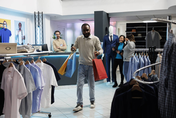 Boldog bolti mániákus, aki ruhadarabokkal teli papírzacskókat cipel a ruhaboltban. Izgalmas afro-amerikai férfi kezében csomagok, miközben vásárol és vásárol a bevásárlóközpont butikban - Fotó, kép